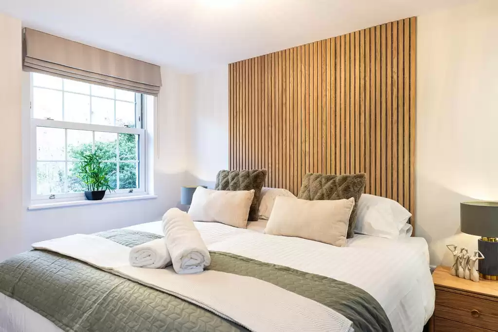 A bed with white soft furnishings, complemented by a green blanket at the bottom of the bed and matching cushions. Behind the bed is a section of wood paneling across the wall. Over the window hangs a beige blind.