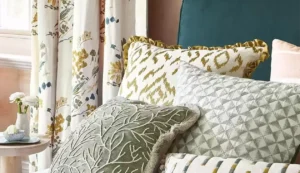 A bed with five upholstered pillows placed on it. Behind the bed hang a pair of patterned curtains.