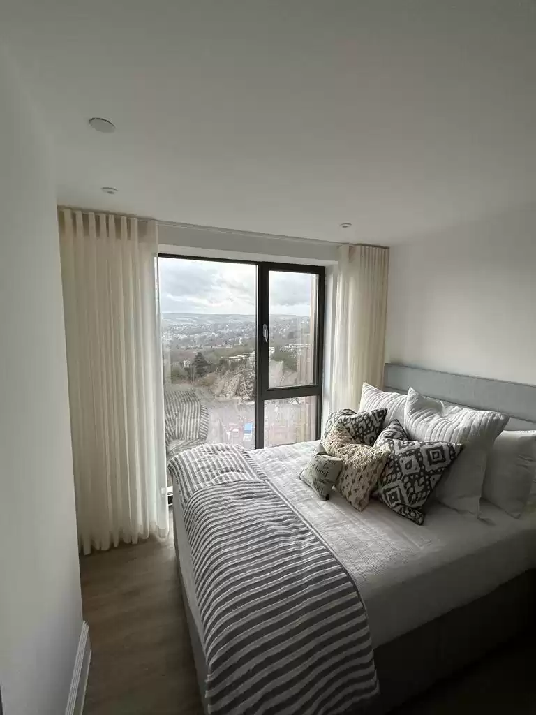 A room with a double bed next to a window covered by sheer white curtains.